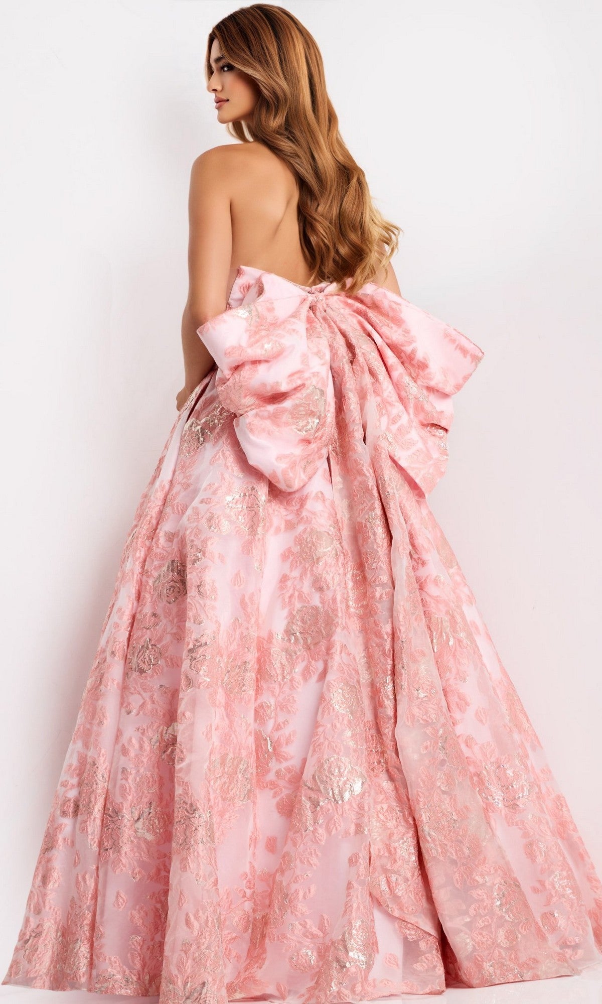 A woman wearing the Jovani 47702 long prom dress—a strapless jacquard ball gown with a pink floral pattern and a large back bow—stands against a plain white background.
